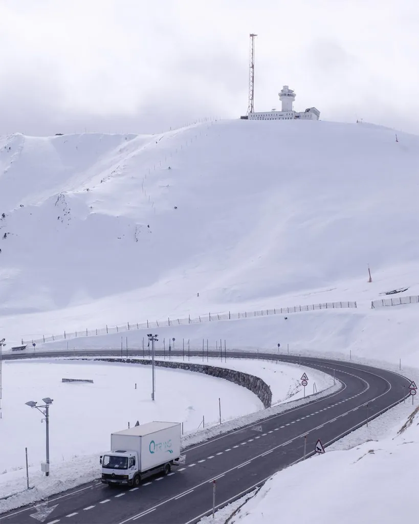 Un camion Q-Trans circule sur un chemin enneigé au pied d'une montagne.